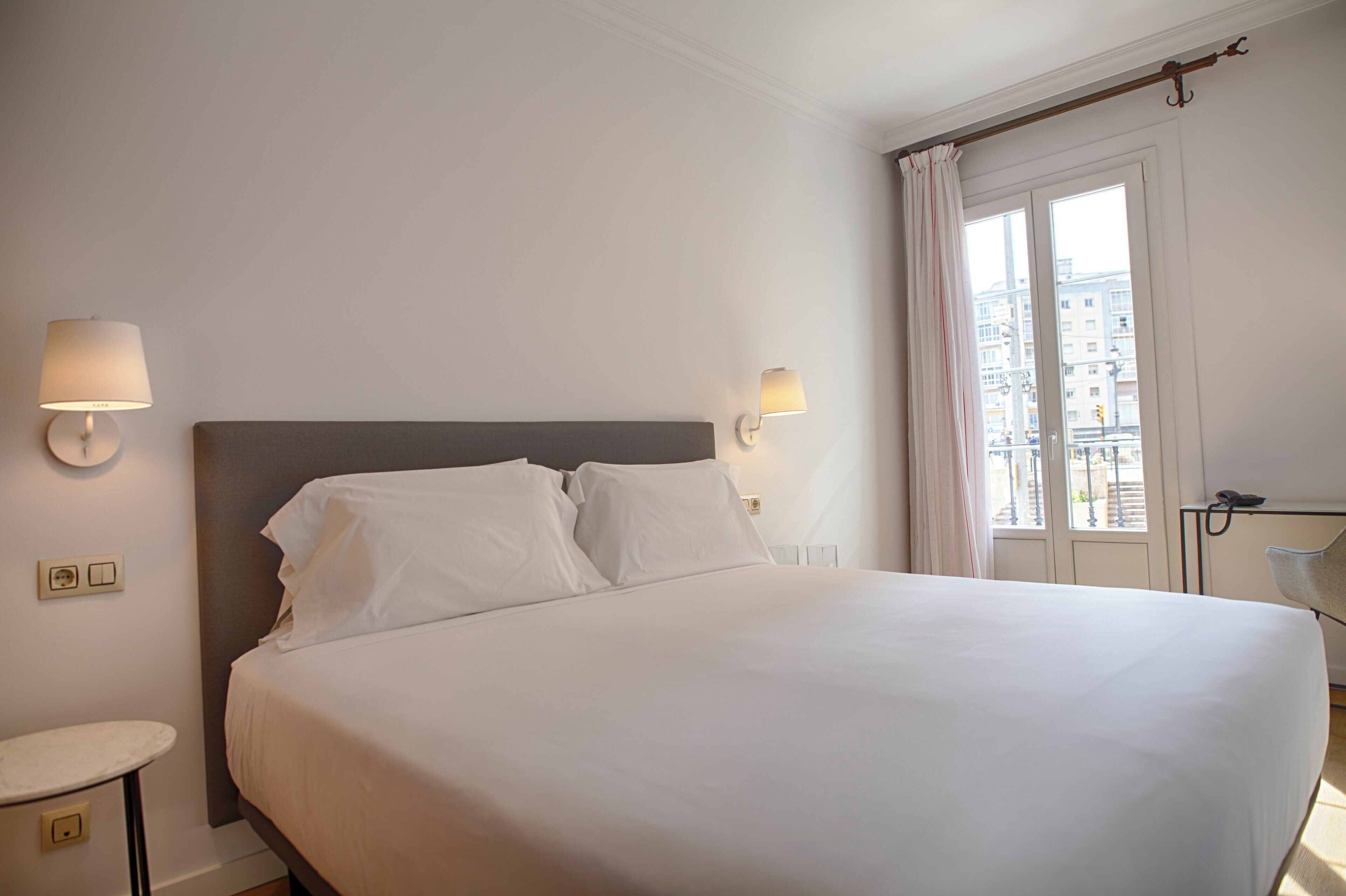 A hotel room with a large bed, bedside lamps, a nightstand, a chair, and a window with curtains.