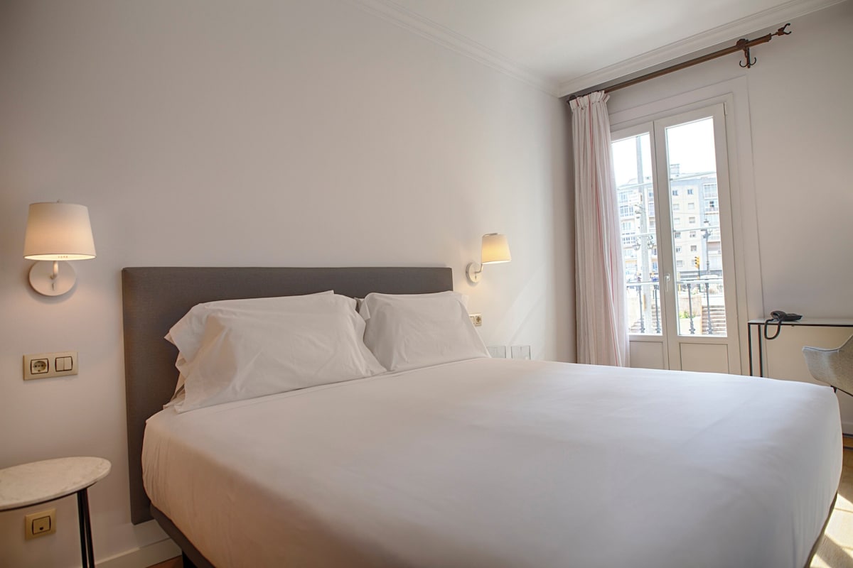 Habitació d'hotel amb un llit gran, làmpades de nit, una taula de nit, una cadira i una finestra amb cortines.