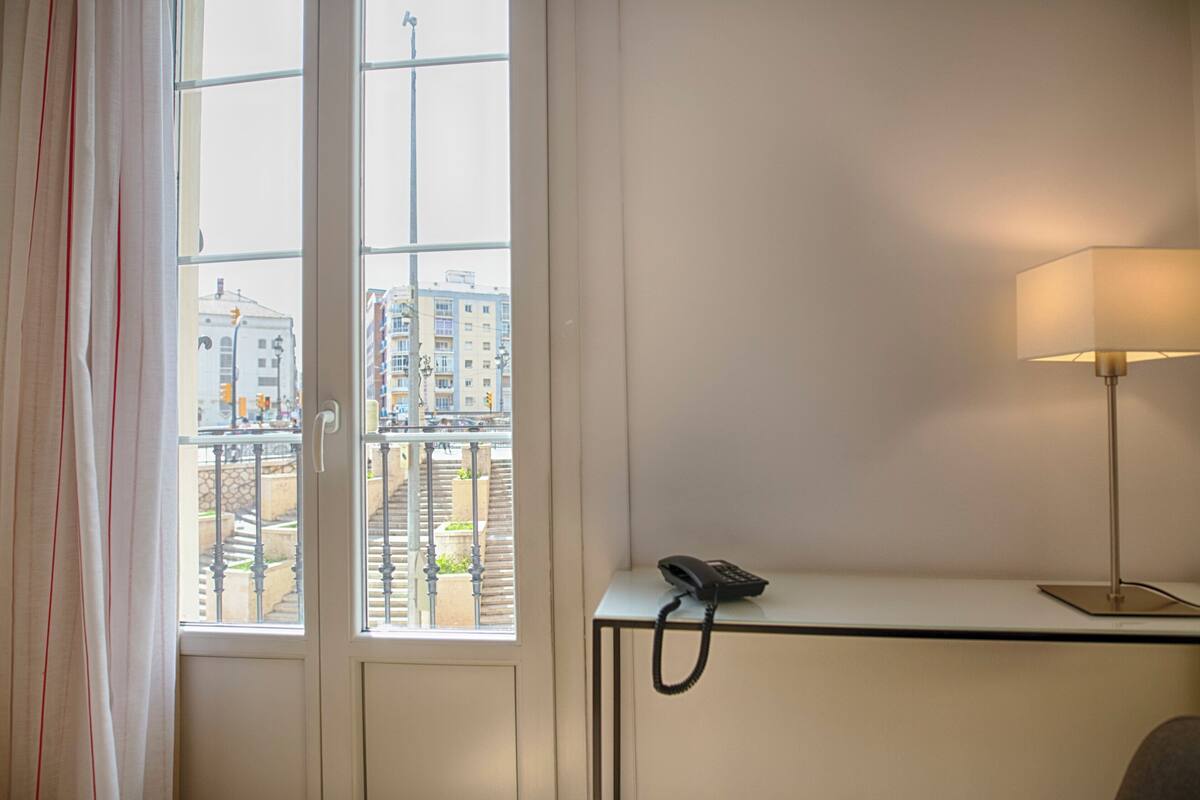 Habitación de hotel con escritorio, teléfono y ventana con vistas a edificios y una escalera.