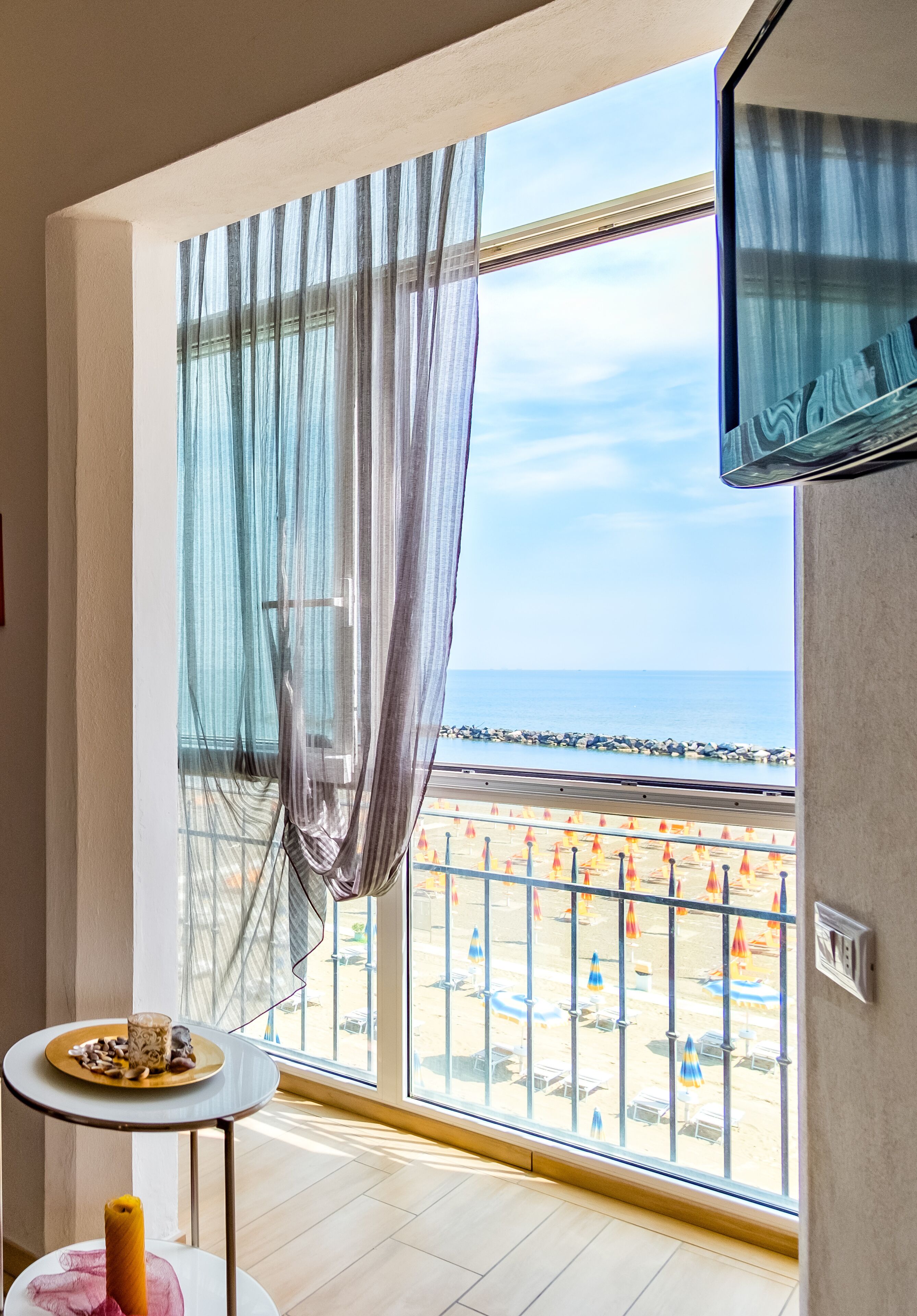 Una habitación con un ventanal grande con vistas a la playa, una mesita con un plato y una botella, y un televisor montado en la pared.