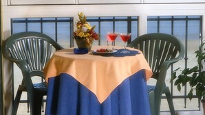 A table with a tablecloth, two chairs, a vase with flowers, and two red drinks on a balcony with a view of the sea.