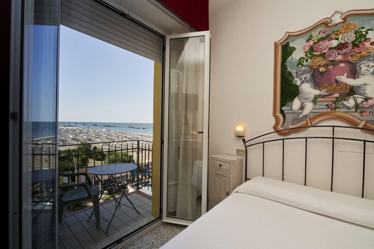 A balcony with a table and chairs, a bed, and a painting of cherubs holding flowers.