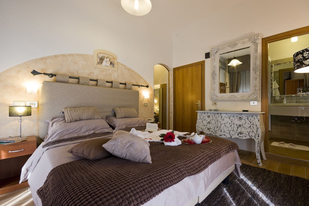 A neatly made bed with a brown blanket, a bedside table with a lamp, a mirror, and a bathroom with a shower and a towel rack.