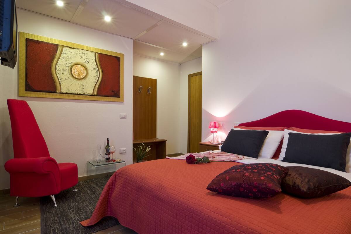 A hotel room with a red bed, a red armchair, a painting on the wall, and a wooden door.