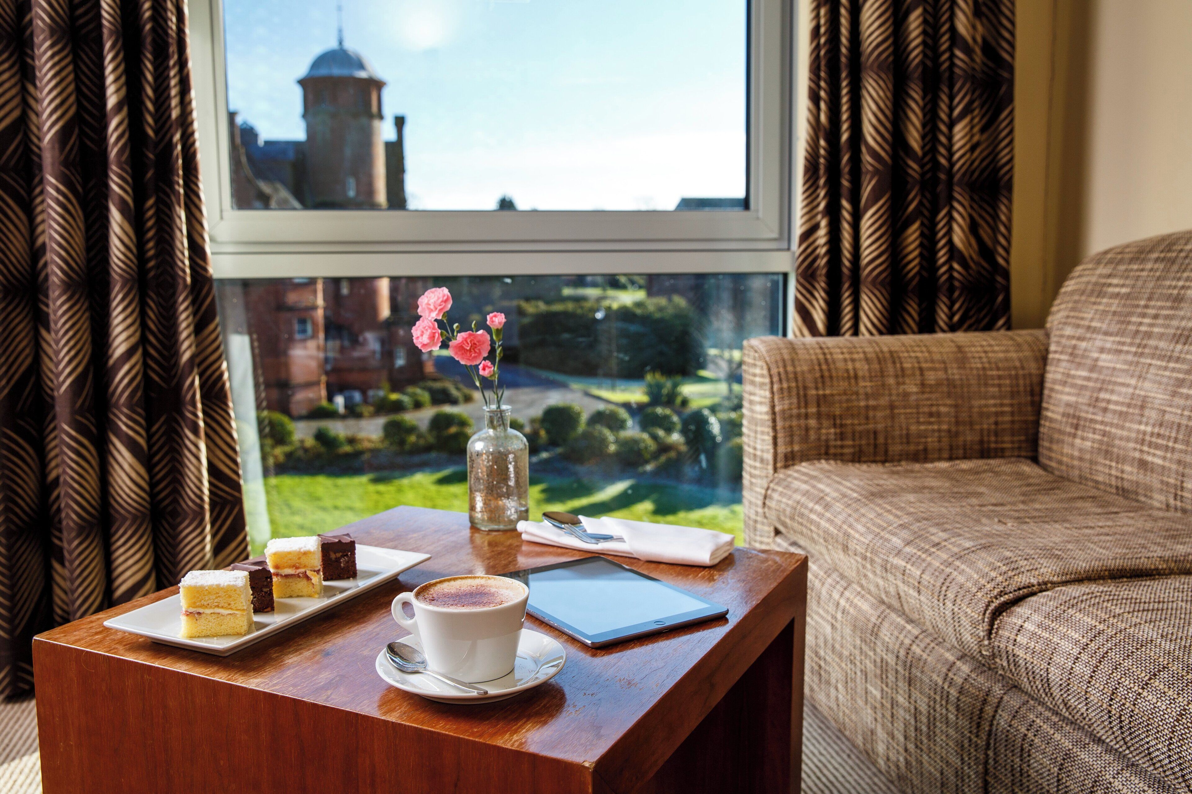 A room with a view of a tower through a window, a wooden table with a cup of coffee, a vase with flowers, and a plate with desserts.