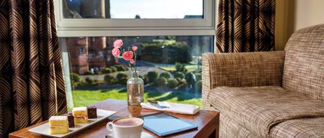 A room with a view of a tower through a window, a wooden table with a cup of coffee, a vase with flowers, and a plate with desserts.