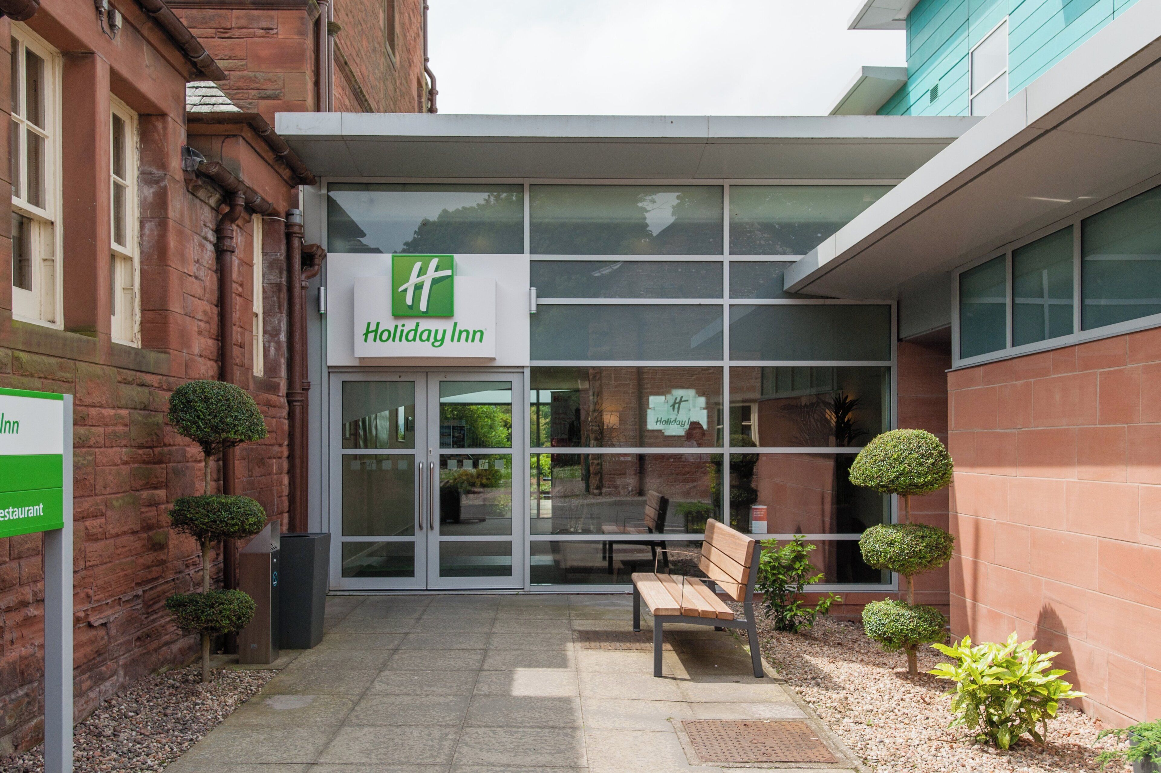 A Holiday Inn hotel entrance with a glass door, a bench outside, and a small garden area with trimmed bushes.