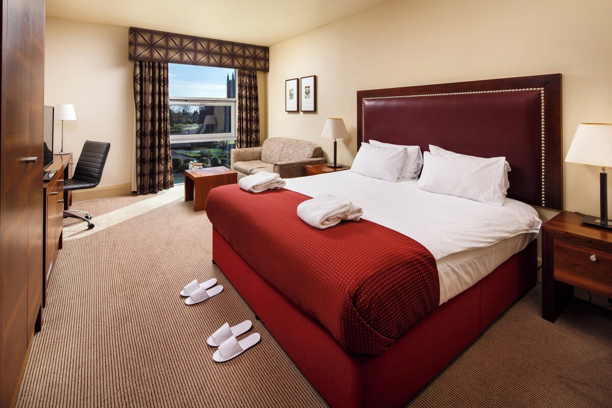 A hotel room with a large bed, a desk with a chair, a sofa, and a window with curtains.