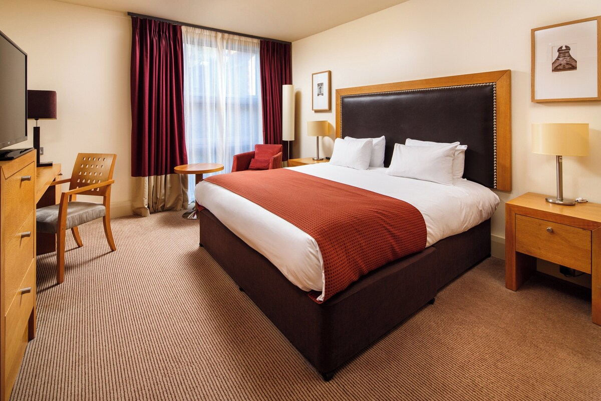 A hotel room with a large bed, a TV, a desk, a chair, and a window with curtains.