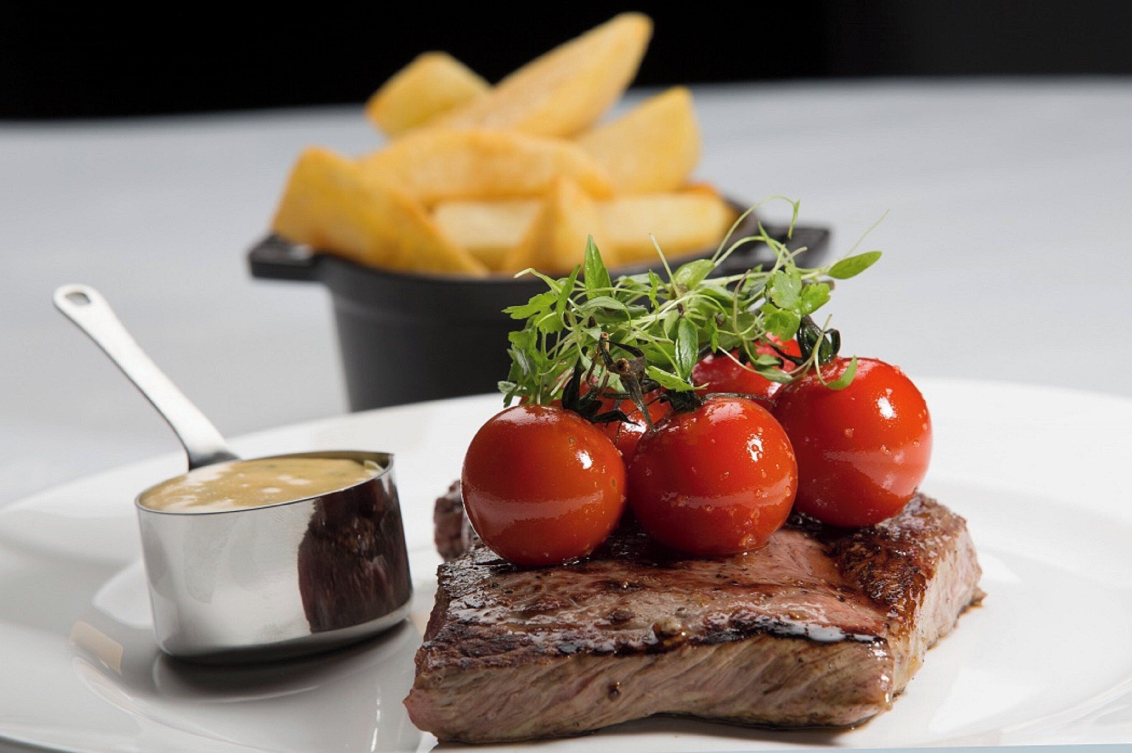 A plate with a steak, cherry tomatoes, and a small bowl of sauce.