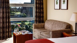 A hotel room with a large window, a sofa, a bedside table with a lamp, and two framed pictures on the wall.