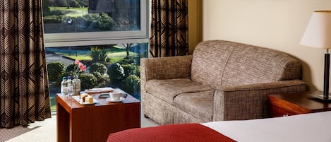 A hotel room with a large window, a sofa, a bedside table with a lamp, and two framed pictures on the wall.