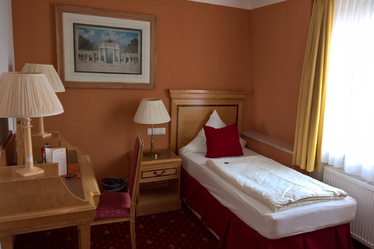 A hotel room with a single bed, a desk with a lamp, a chair, a framed picture on the wall, and a window with curtains.
