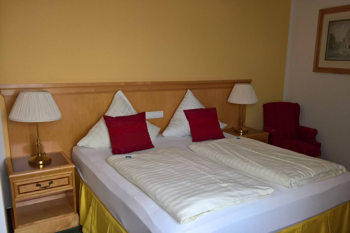 A double bed with two pillows, a nightstand with a lamp, a chair, and a small table with a drawer.