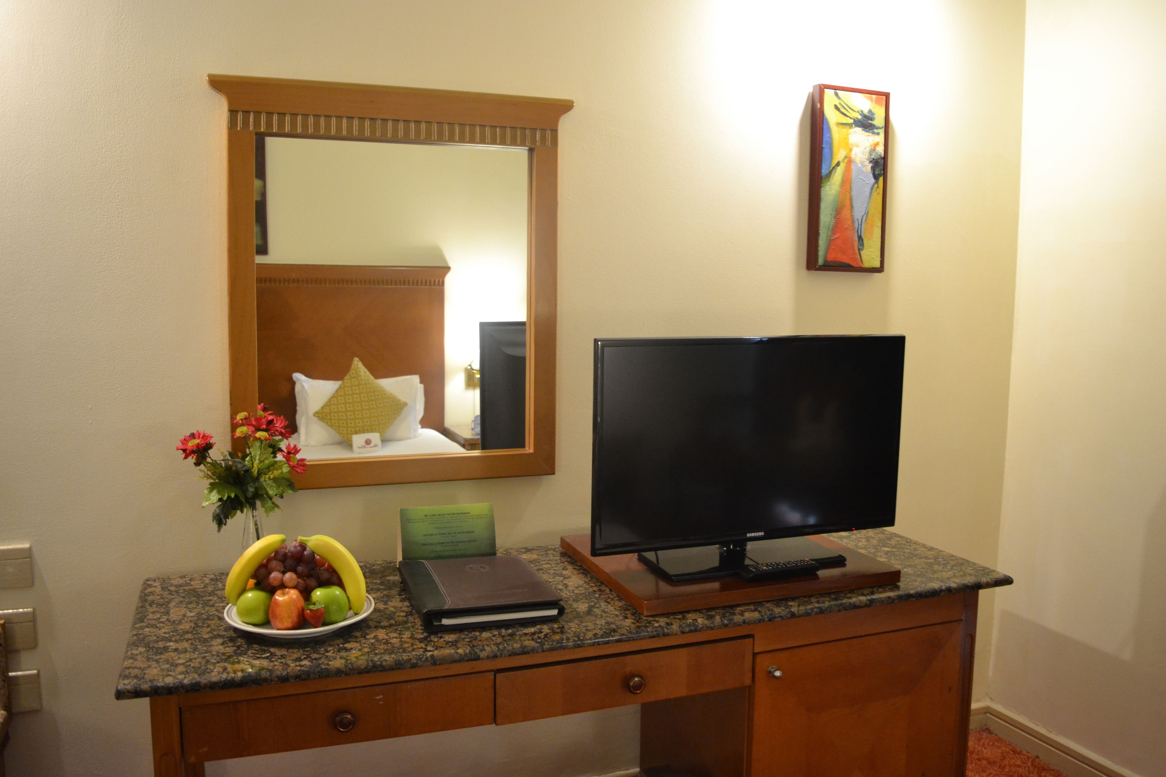 Escritorio de habitación de hotel con una televisión de pantalla plana, un frutero, un florero con flores, un libro y un espejo sobre el escritorio.