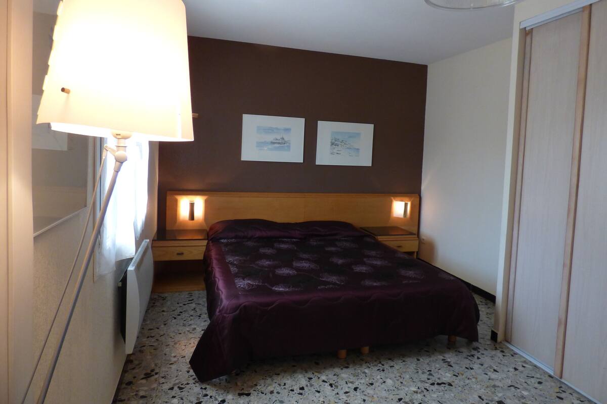 Un dormitorio pequeño y acogedor con cama, lámparas de noche y un radiador. Hay dos cuadros en la pared y un armario a la derecha.
