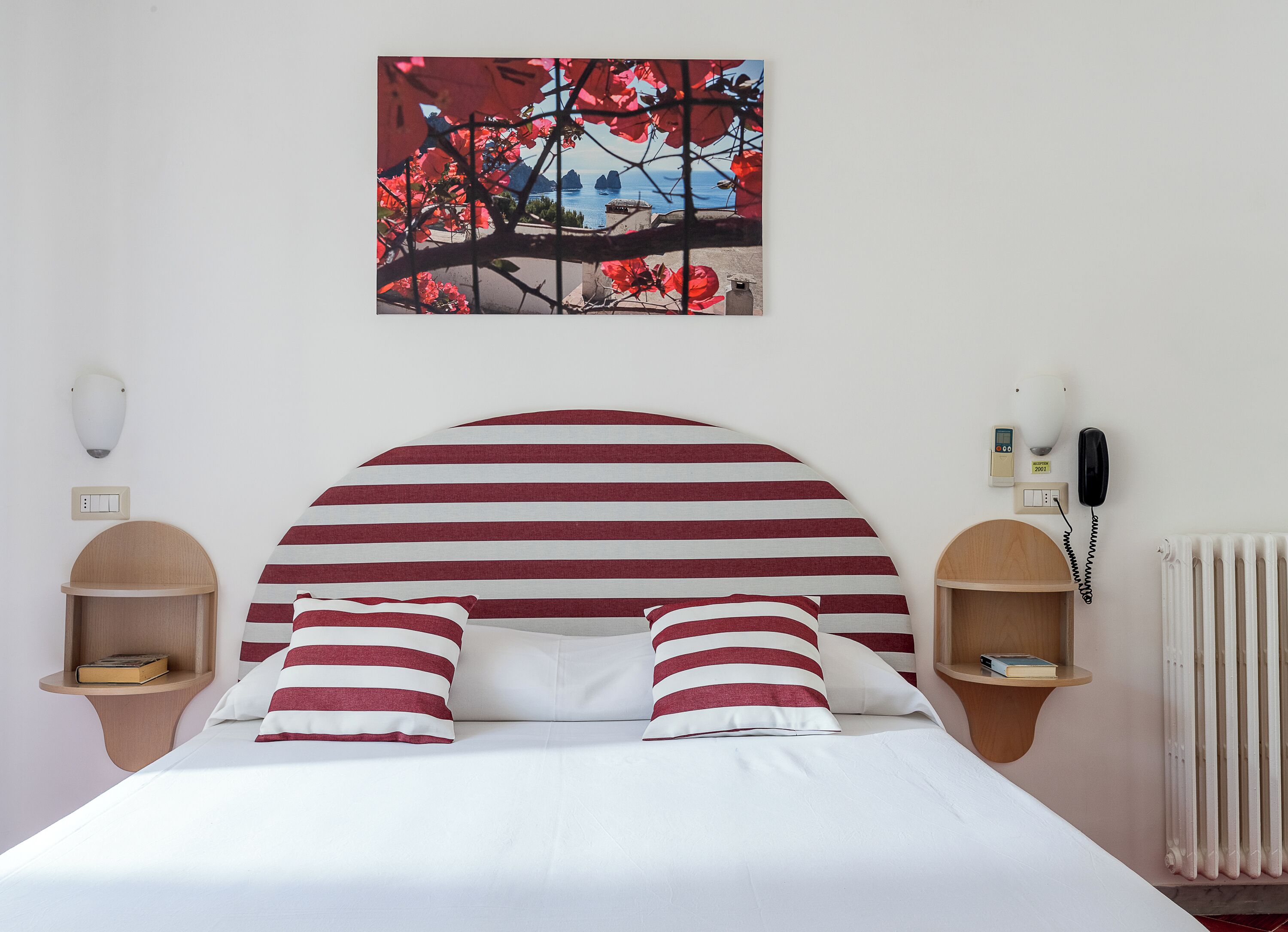 A bed with a striped headboard, two bedside tables with books, a wall-mounted phone, and a radiator.