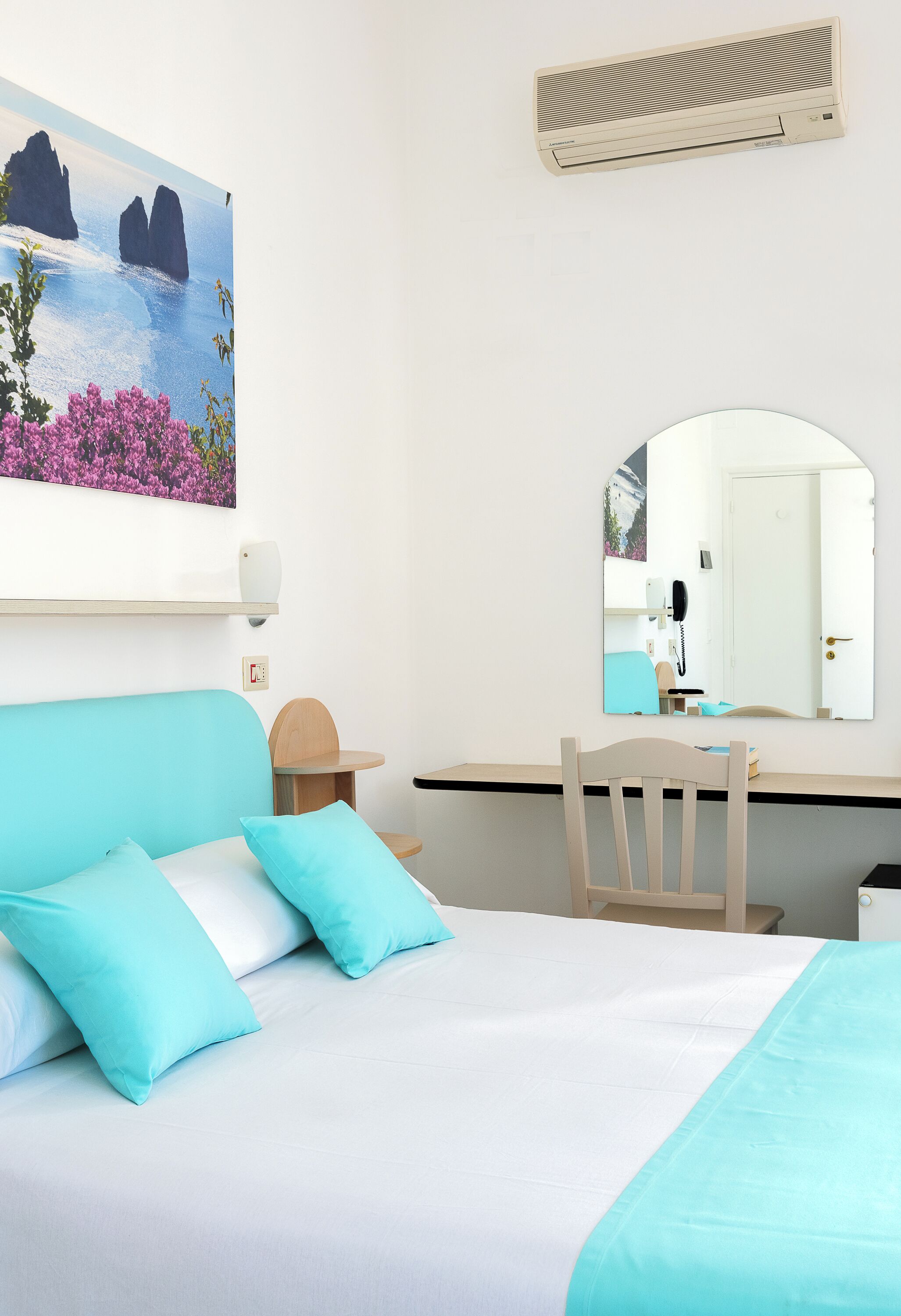 Une chambre d’hôtel comprenant un lit, un bureau, une chaise, un miroir et un tableau représentant un paysage côtier.