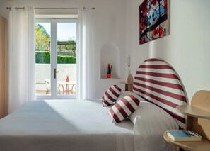 A hotel room with a bed, a view of a garden through a glass door, a small table with a book, and a painting on the wall.