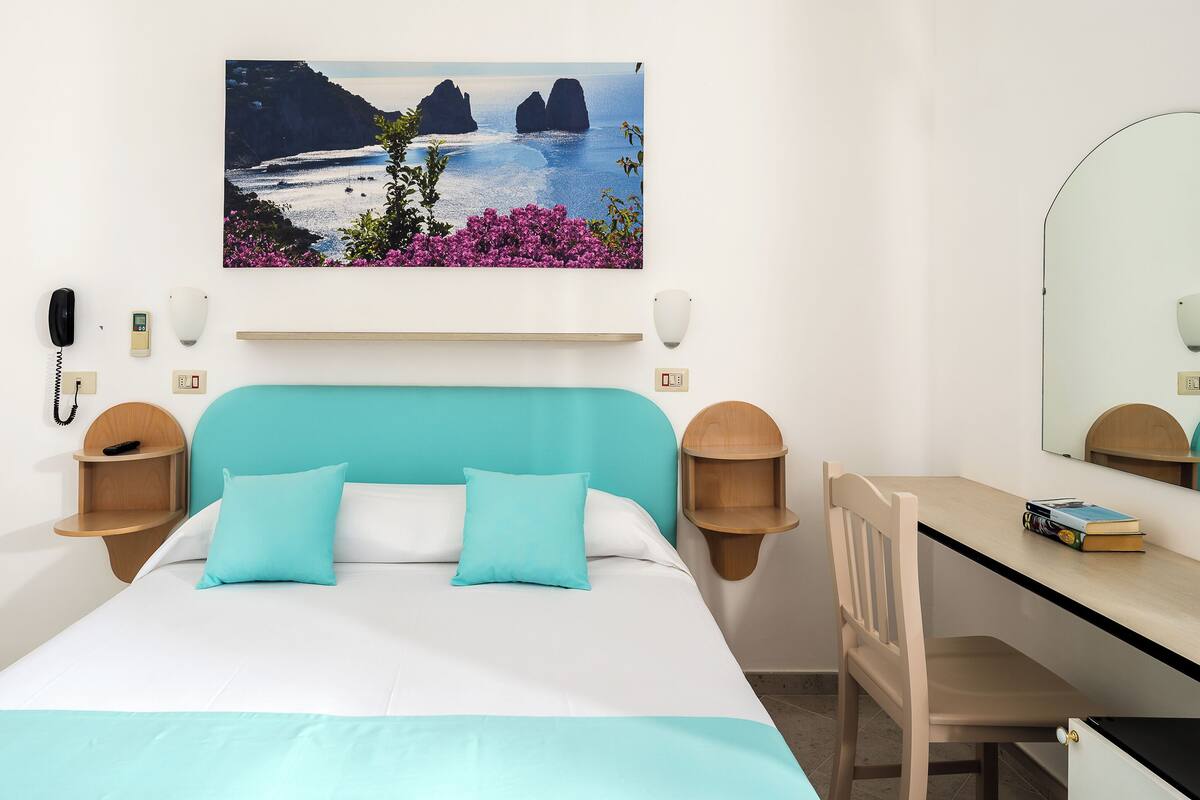 A hotel room with a bed, a desk, a chair, a phone, a lamp, and a painting of a coastal landscape.