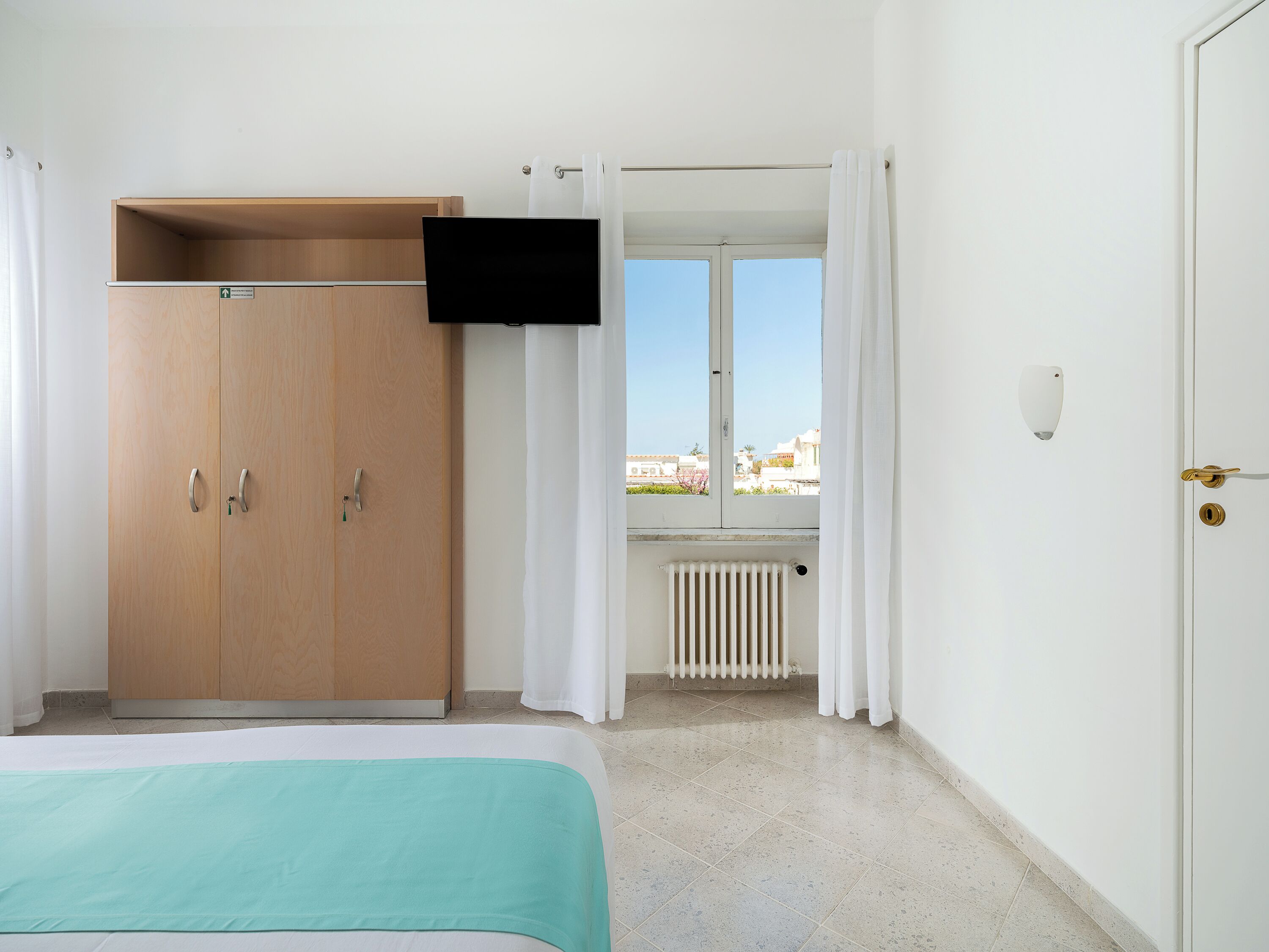 Un dormitorio con cama, armario, televisor, ventana con cortinas y radiador.