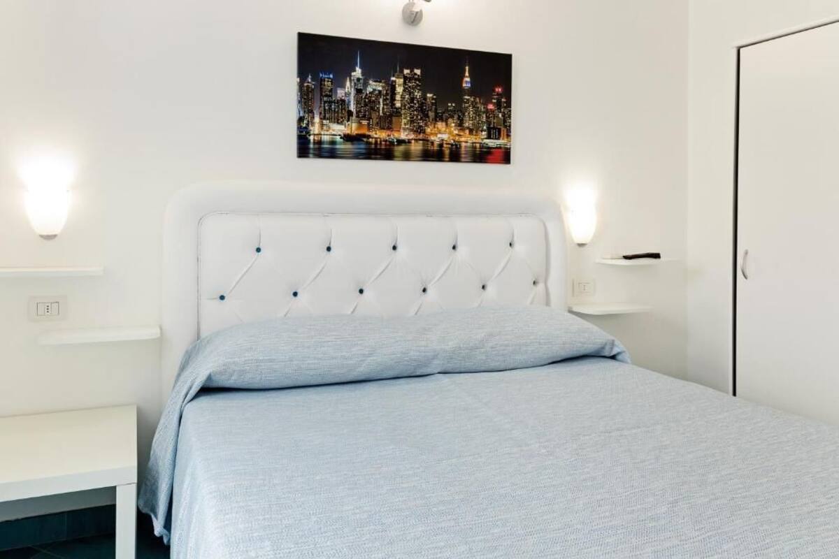 A bedroom with a white headboard, a bed with a light blue bedspread, two bedside tables, a nightstand, a wall-mounted light, and a cityscape picture on the wall.