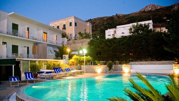 Piscina com espreguiçadeiras, um muro de pedra e edifícios ao fundo.