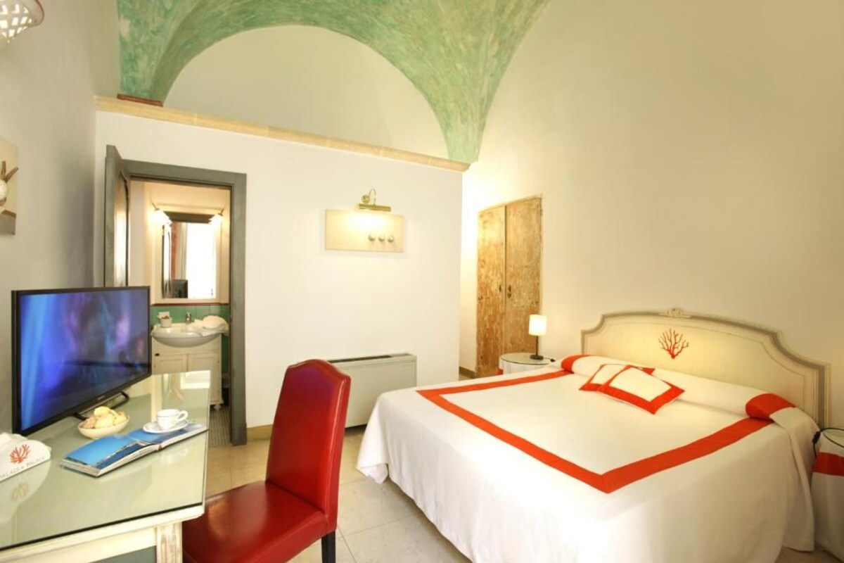A hotel room with a bed, a desk with a computer, a red chair, a TV, and a bathroom with a sink and mirror.