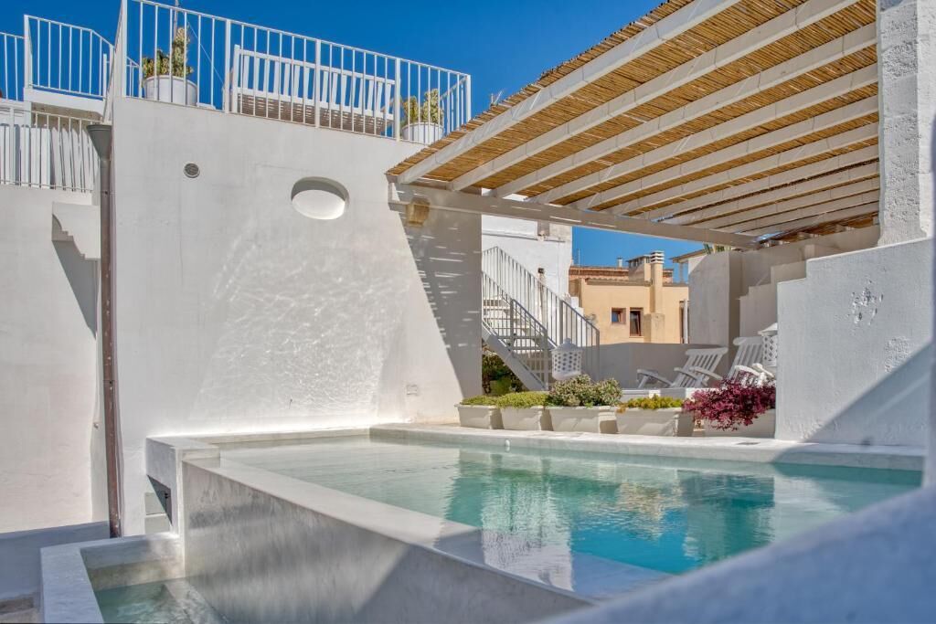 A modern outdoor pool area with a pergola, white walls, and a staircase leading to a rooftop terrace.