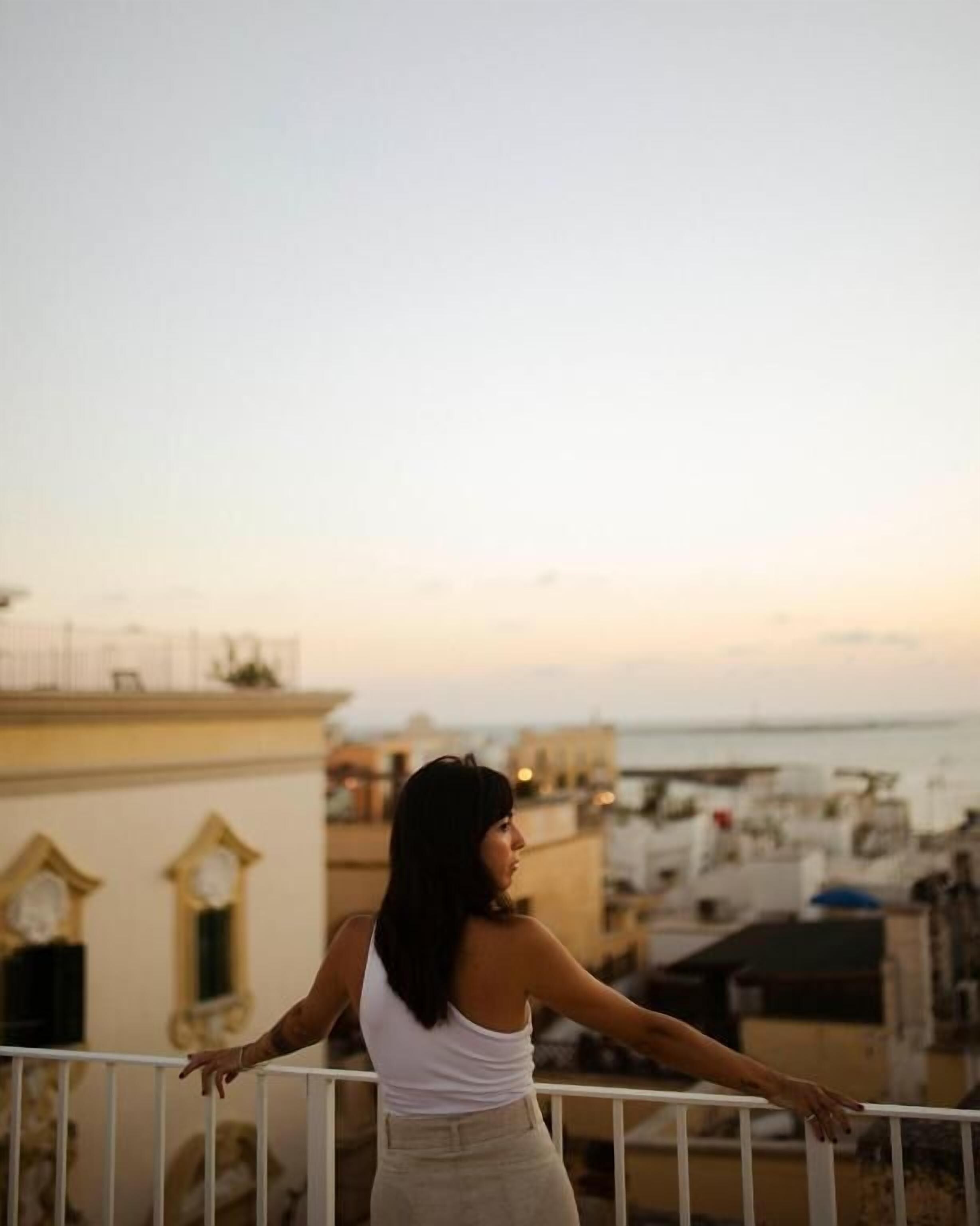 A person standing on a balcony with a railing, overlooking a cityscape with buildings and a body of water.