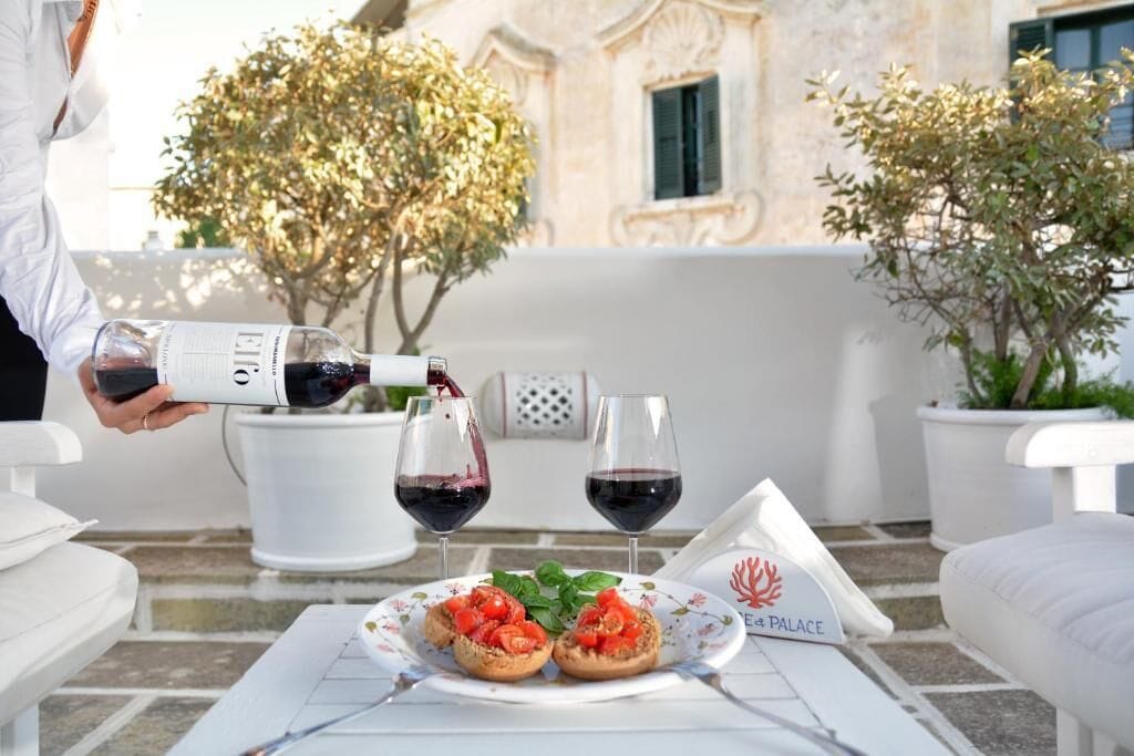 Une table avec une assiette de bruschettas et deux verres de vin rouge, une personne versant du vin, et un bâtiment en arrière-plan.