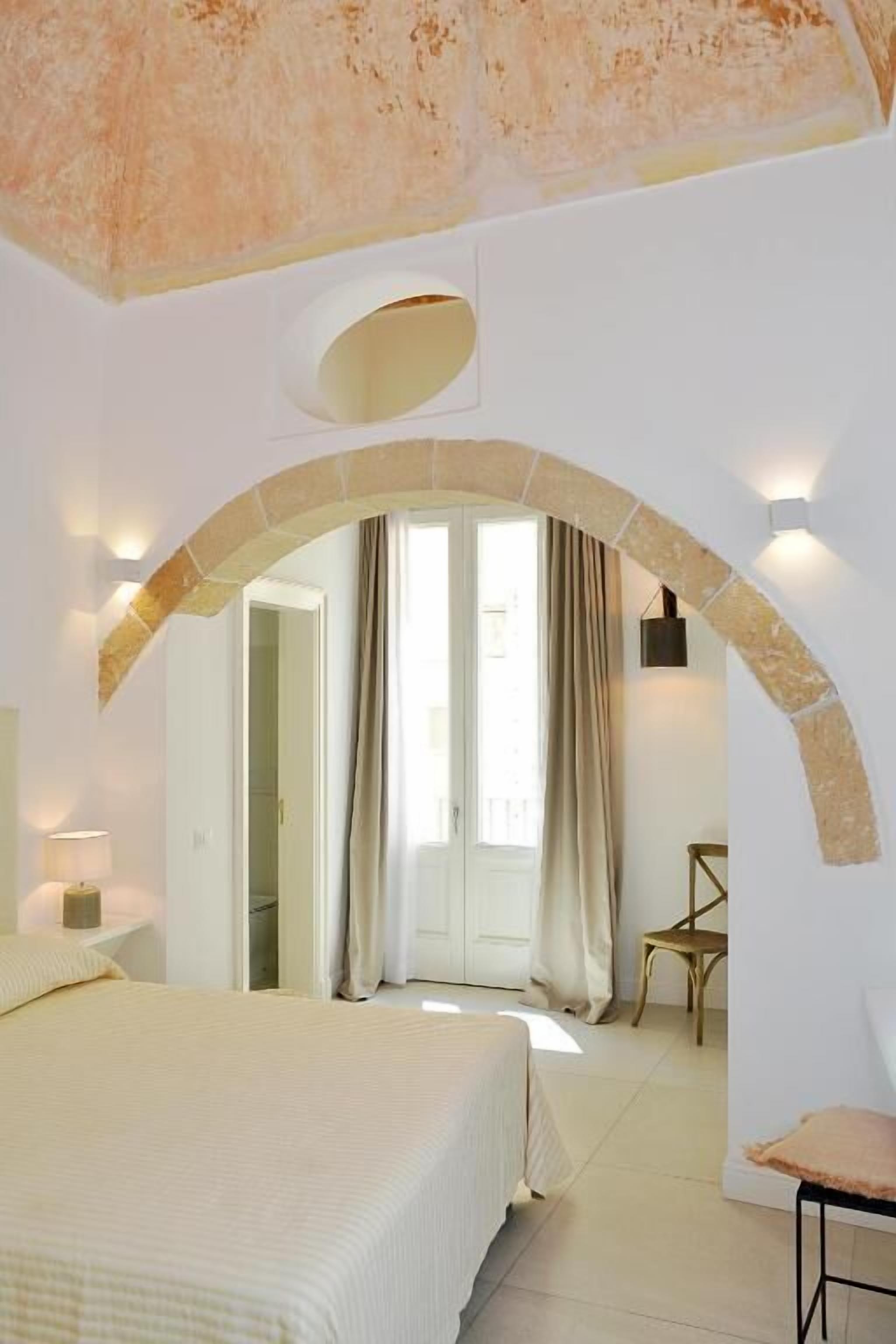 Un dormitorio con cama, una silla, un arco de entrada y una ventana con cortinas.