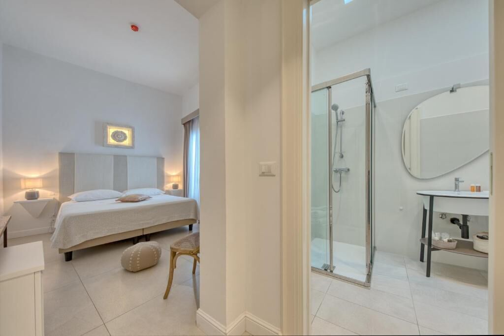 A modern bedroom with a bed, bedside table, and a small stool. Adjacent is a bathroom with a shower, sink, and mirror.