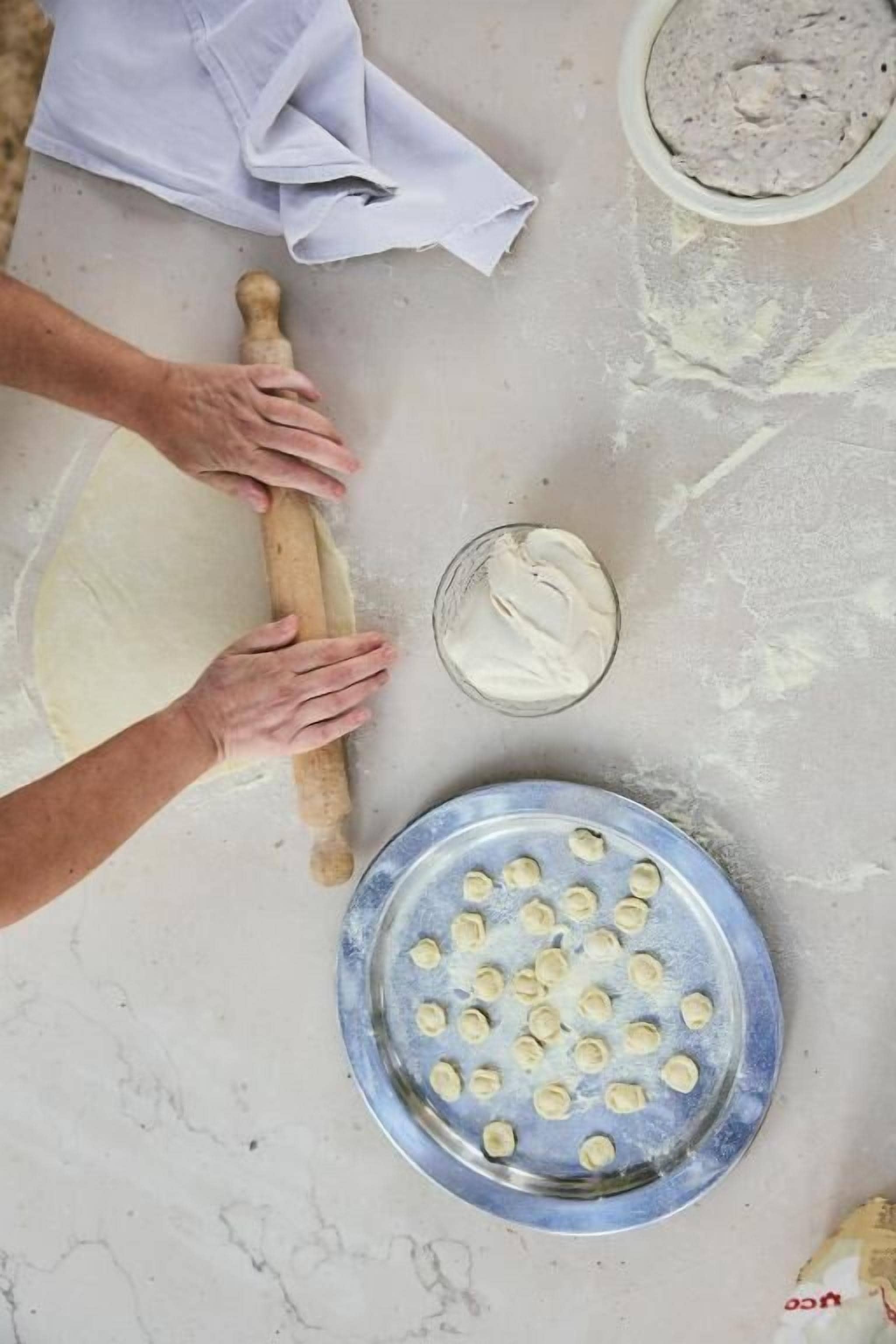 Una persona extiende masa sobre una encimera de mármol, usando un rodillo, un bol de relleno y un plato con porciones de masa ya formadas.