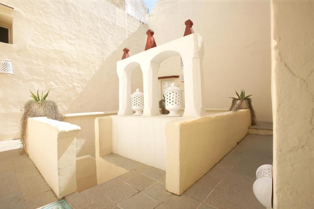 Un patio bañado por el sol, con una fuente blanca, plantas en macetas y suelo de baldosas.