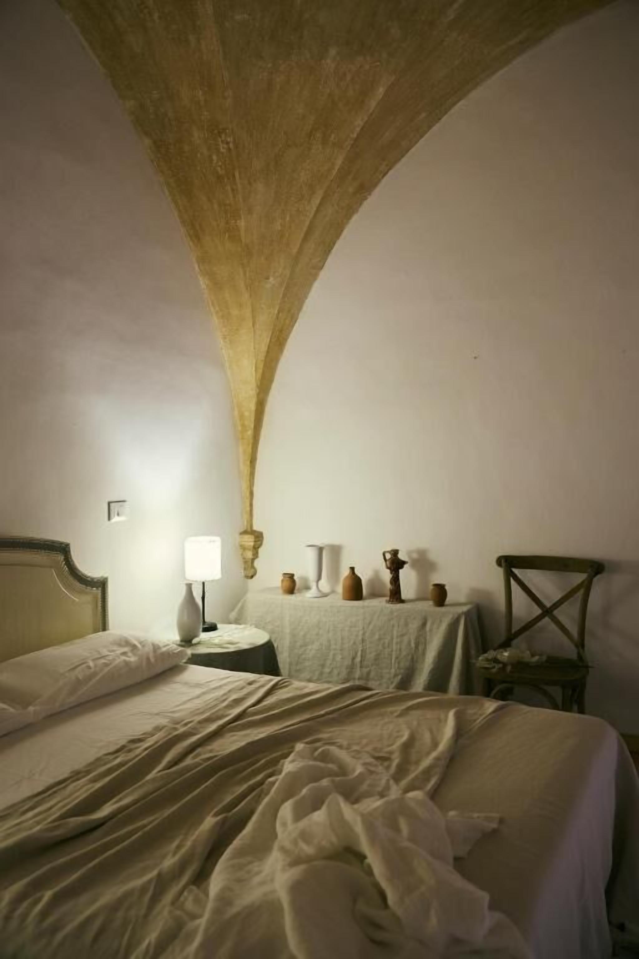 A bedroom with a large arched ceiling, a bed with white linens, a bedside table with a lamp, and a small table with decorative items.