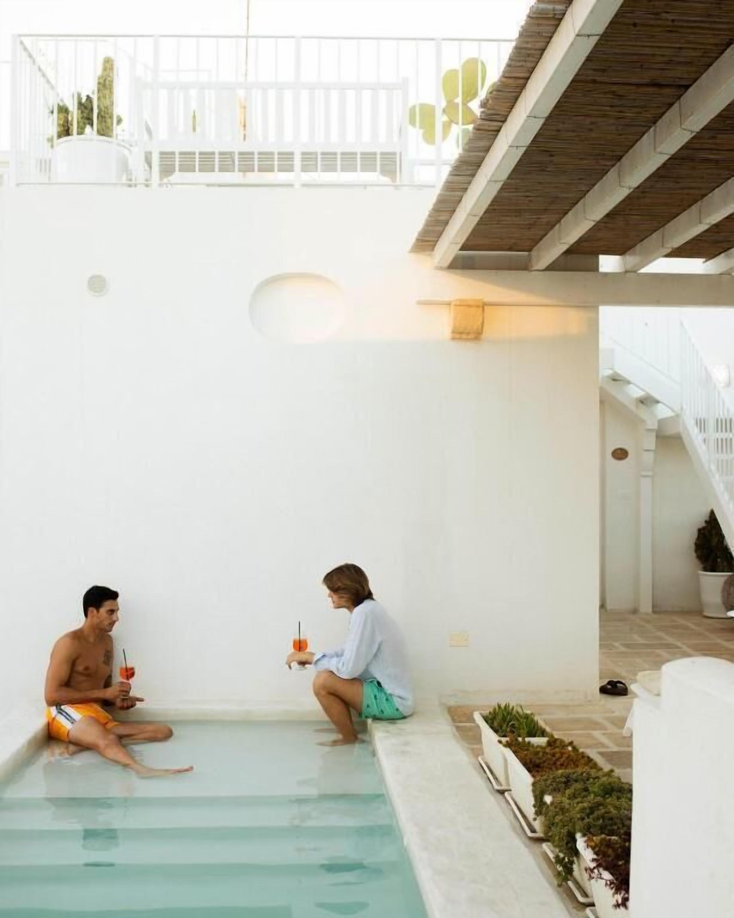 A man and a woman sitting by a pool with a white wall and a balcony in the background.