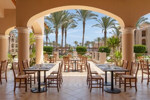 A covered outdoor dining area with tables and chairs, palm trees, and a view of a building with arches.