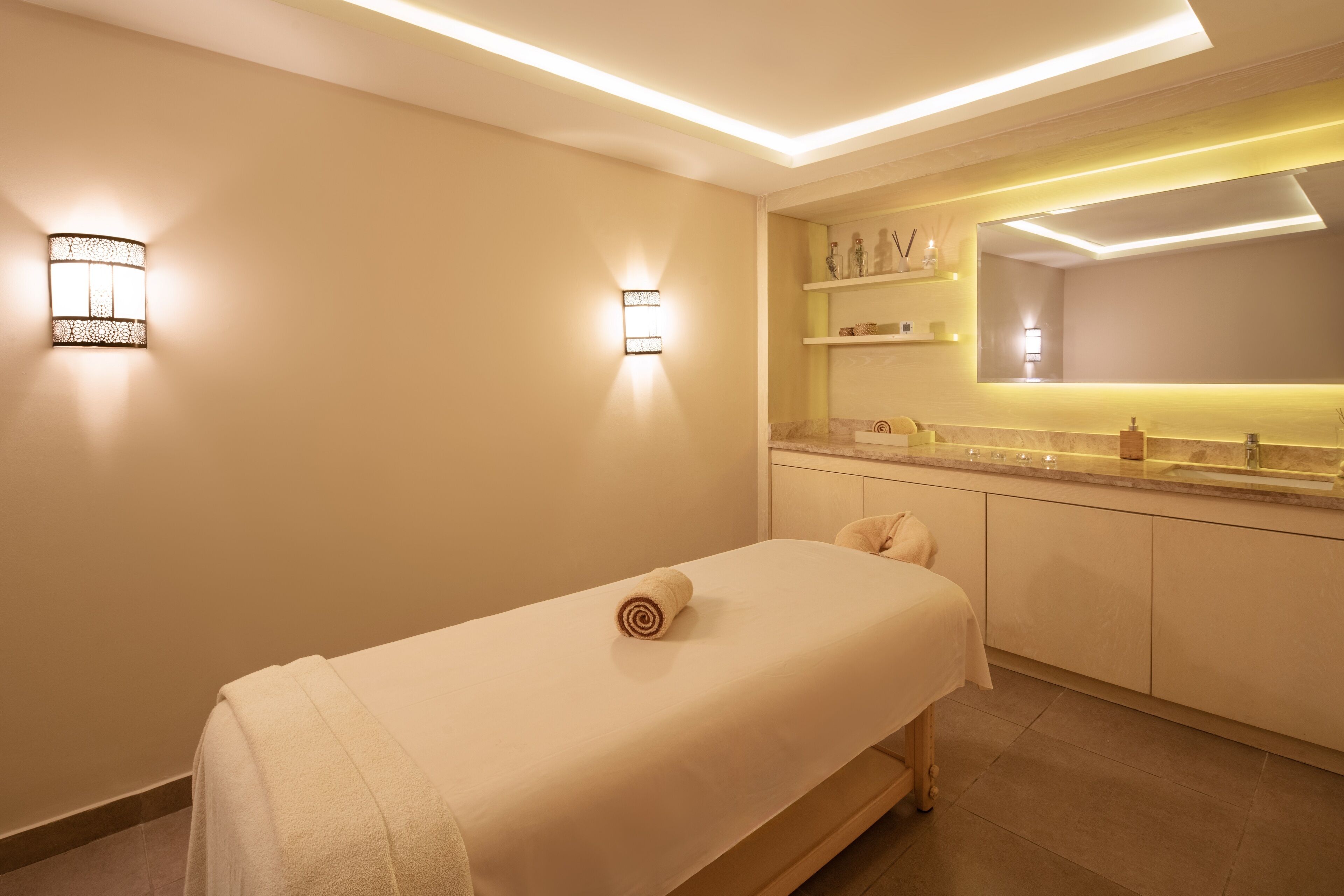 A spa room with a massage table, wall-mounted lights, a vanity with a mirror, and a shelf with toiletries.