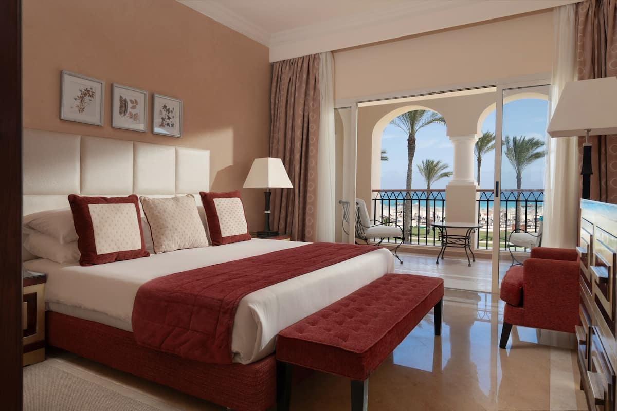 Une chambre d’hôtel dotée d’un grand lit, d’un balcon offrant une vue sur les palmiers et la mer, ainsi que d’un coin repas avec des chaises et une table.