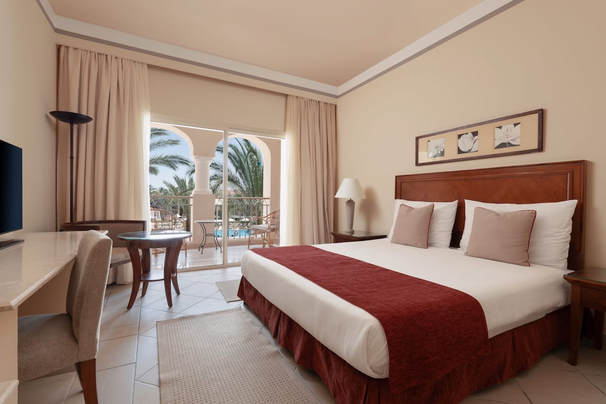 Une chambre d’hôtel avec un grand lit, un bureau, une chaise, une télévision et un balcon offrant une vue sur les palmiers.