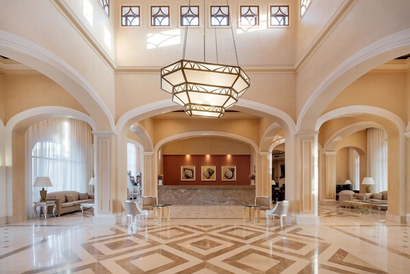 A spacious lobby with a geometric patterned floor, a central chandelier, and a reception desk.