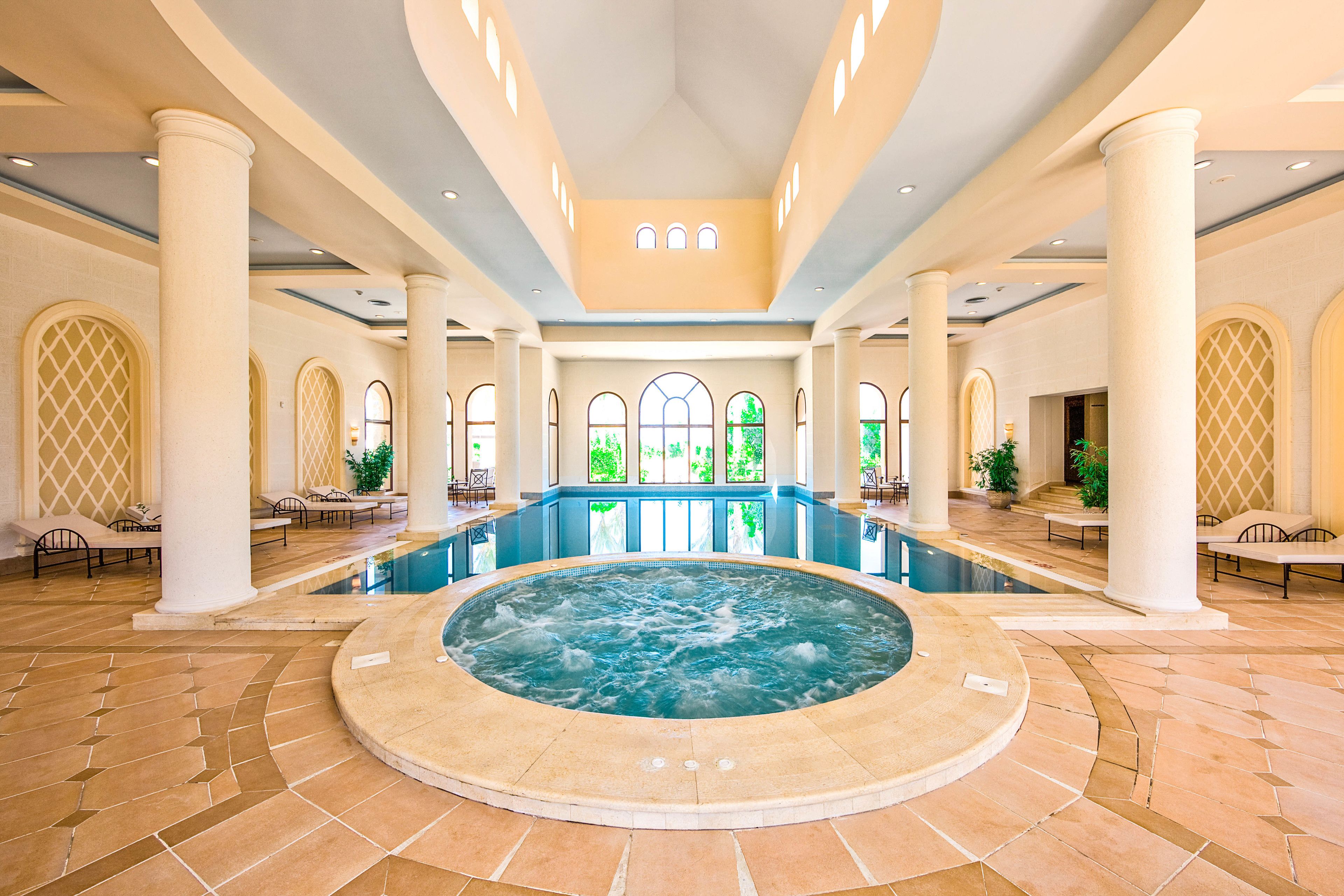 A large indoor swimming pool with a hot tub in the center, surrounded by columns and lounge chairs.