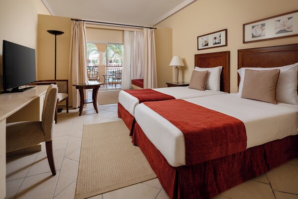 Habitación de hotel con dos camas, un escritorio, una silla, un televisor y un balcón con vistas a palmeras.