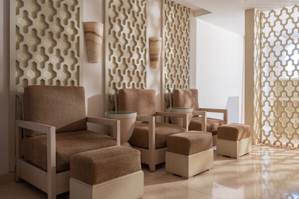 Un spa moderno con paredes color beige estampadas, un sillón grande y acolchado, una mesita y un jarrón decorativo.
