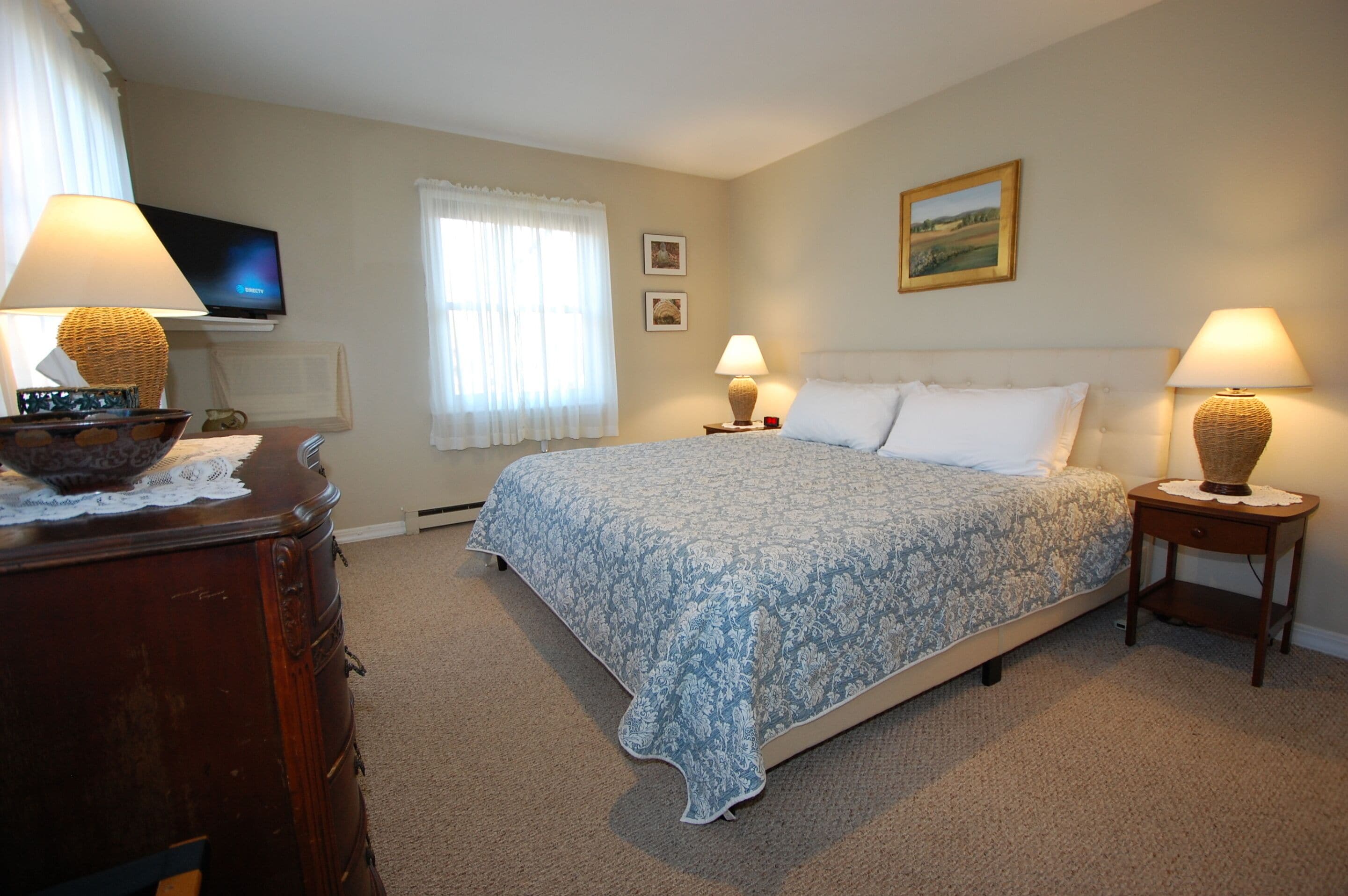 A bedroom with a bed, two bedside tables with lamps, a dresser, a television on the wall, and a window with curtains.