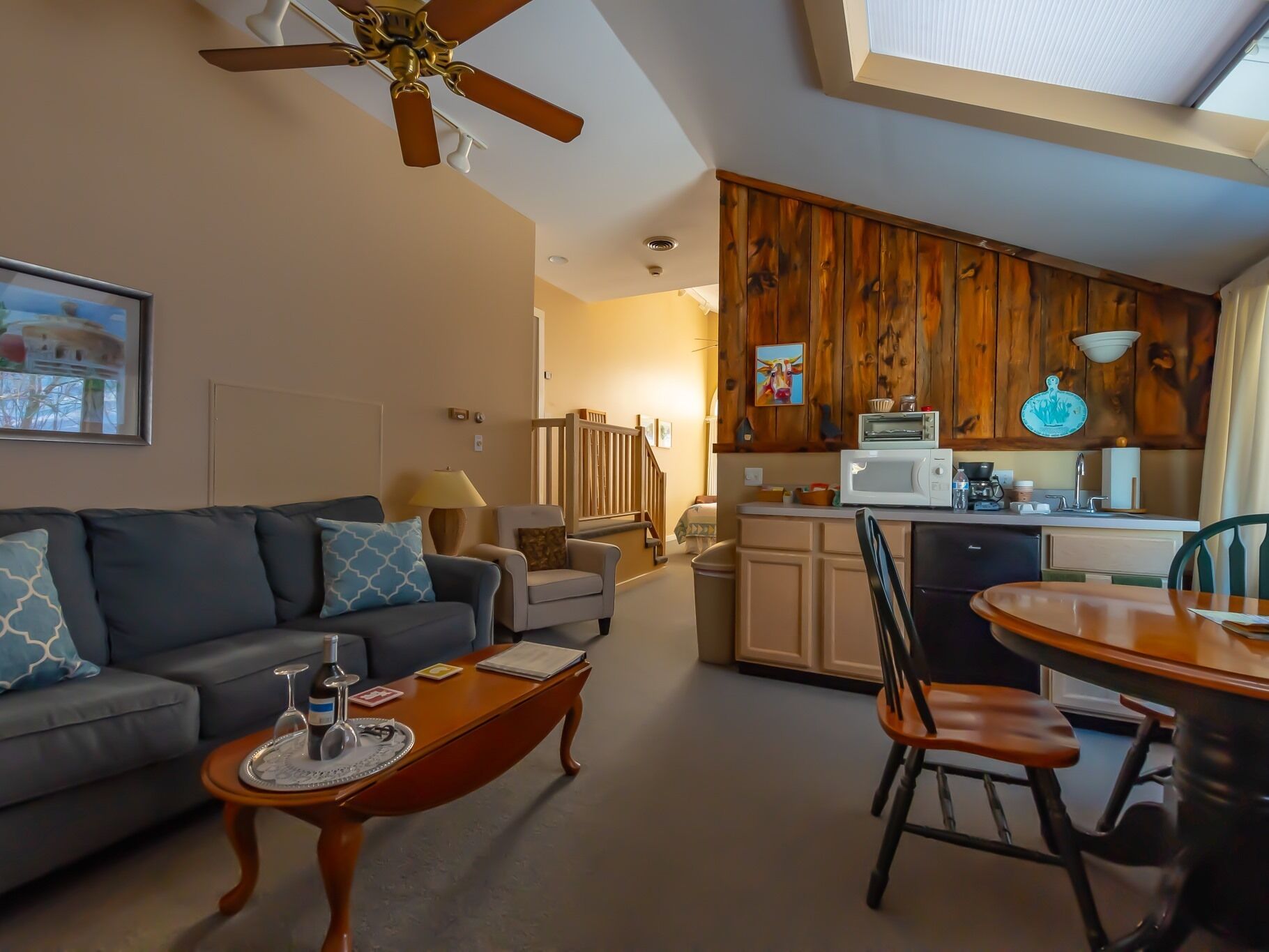 A living room with a sofa, coffee table, and a dining area with a table and chairs. A kitchen with wooden cabinets and a microwave is visible in the background.