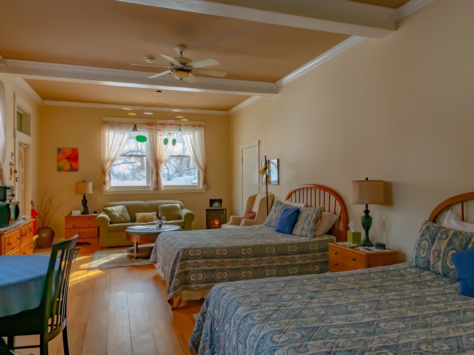 A cozy bedroom with two beds, a ceiling fan, a fireplace, and a view of the outside.
