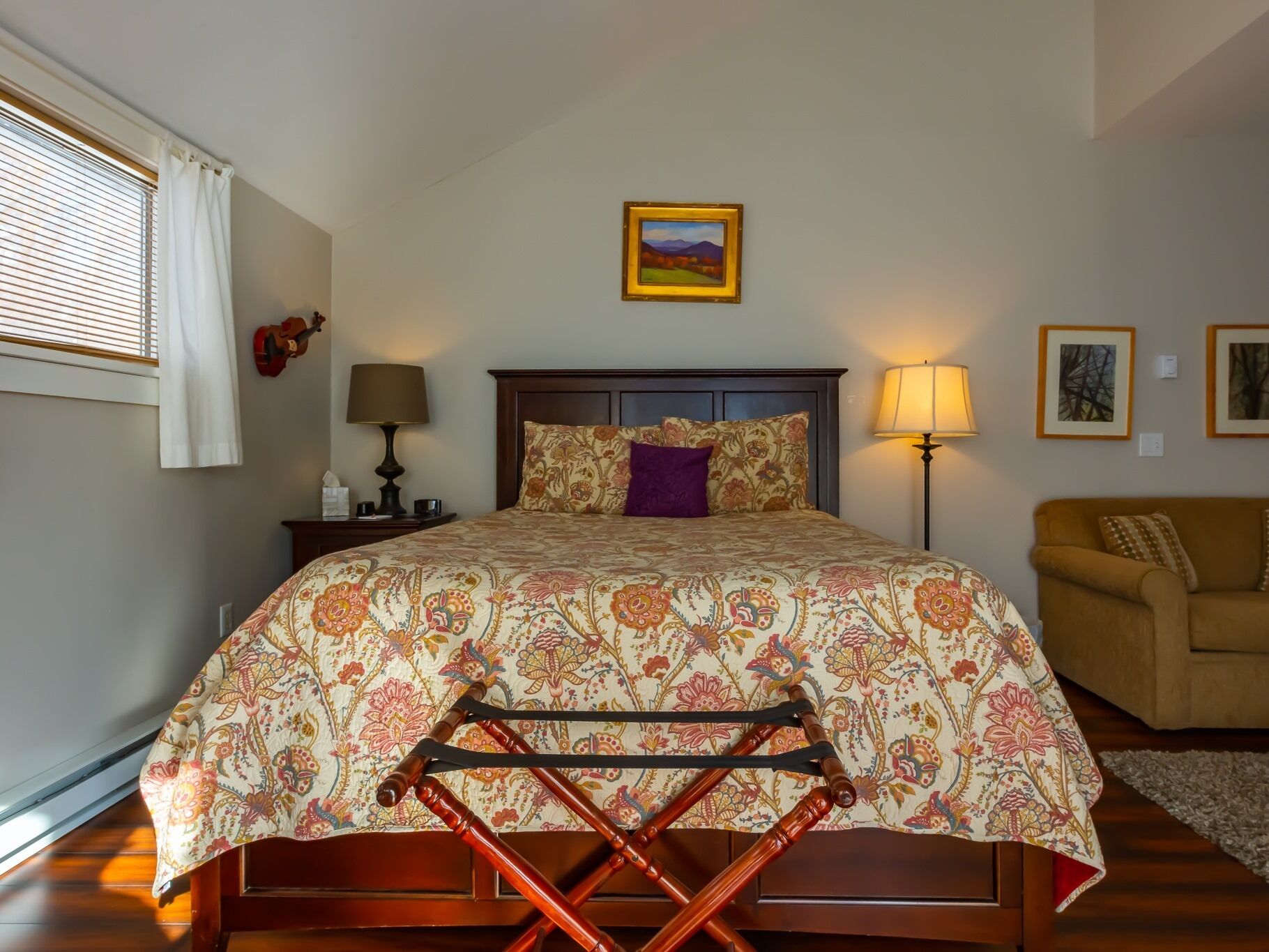 A bedroom with a bed, a nightstand, a lamp, a guitar, a couch, and a window with blinds.