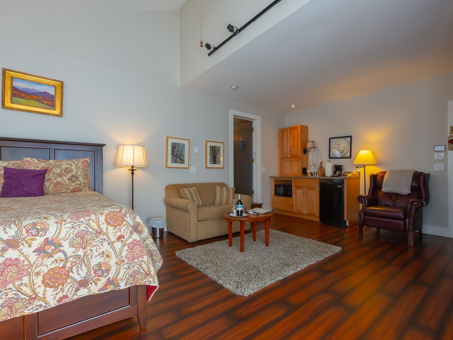 Ein Schlafzimmer mit Bett, Nachttisch, Lampe und Kommode. Ein Wohnbereich mit Sofa, Couchtisch und Sessel ist ebenfalls vorhanden. Man sieht eine Küchenzeile mit Schränken und Kühlschrank.